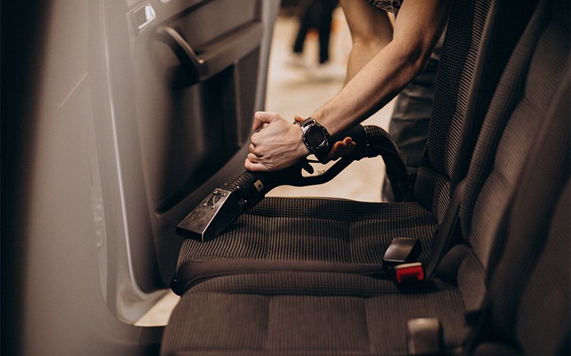 Professional detailing upholstery cleaner vacuuming rental car passenger seat.